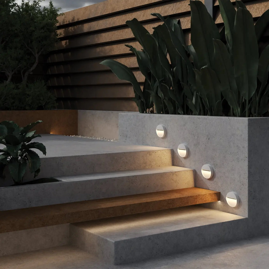 Quatre balises rondes blanches led encastrées diffusant une lumière douce sur un escalier en béton pour une applique extérieure mane
