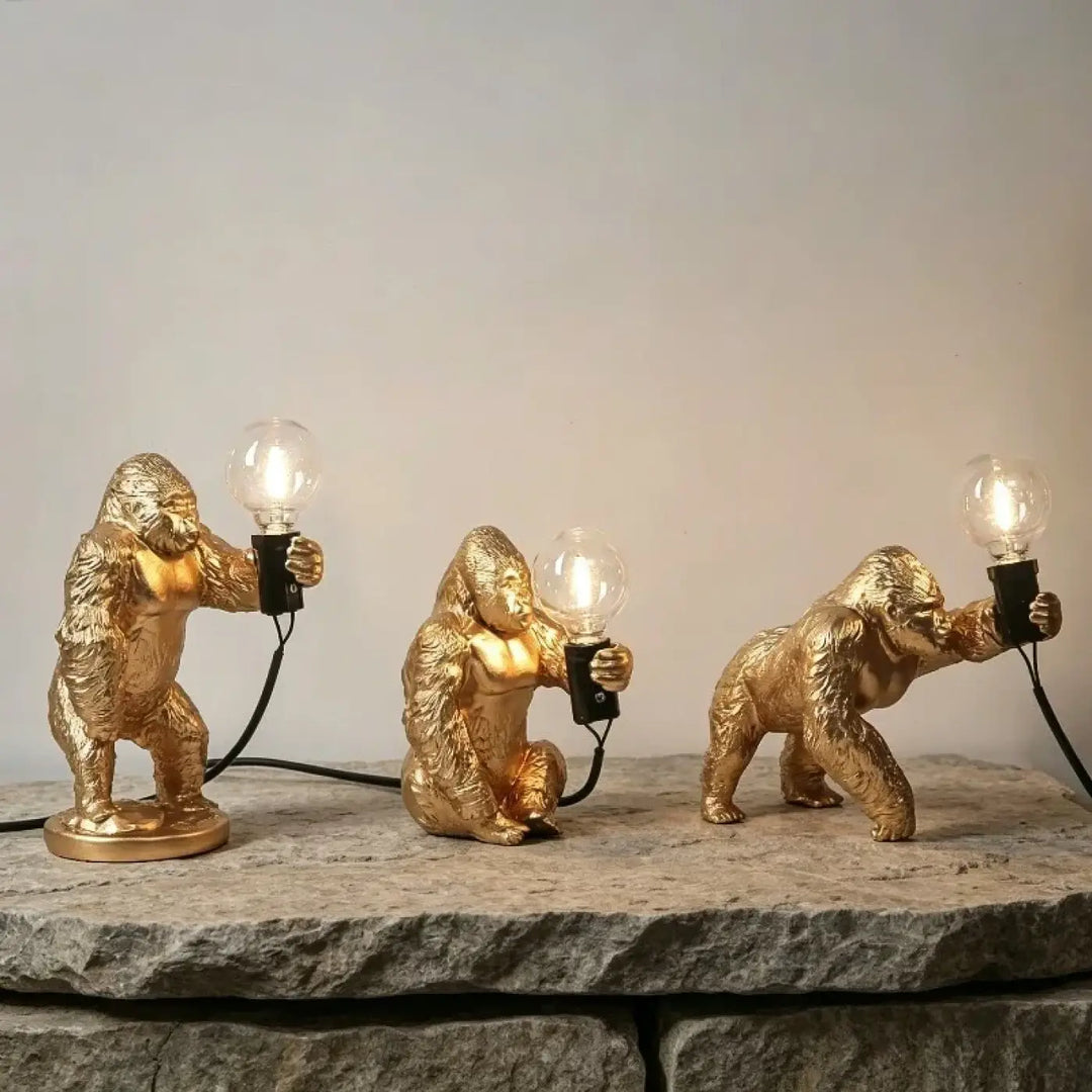 Lampe de table gorenzo avec trois lampes artistiques gorille dorées tenant des ampoules allumées sur pierre