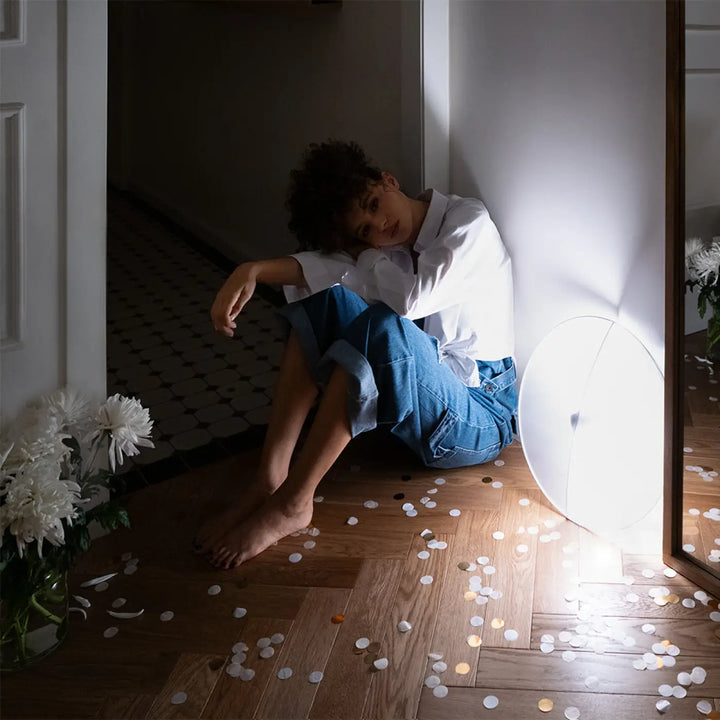 Une personne aux cheveux bouclés assise sur un sol en bois à côté d’une suspension led reflex lumière chaude blanche